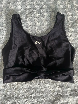 Svart sport-bh från Only Play - Snygg svart sport-bh från Only Play med breda axelband och knutdetalj framtill. Tillverkad i ett glansigt, stretchigt material som ger bra stöd. Logotyp framtill och baktill. Perfekt för träning eller en aktiv vardag. Lappen med storlek är avlklippt men den är en S eller M