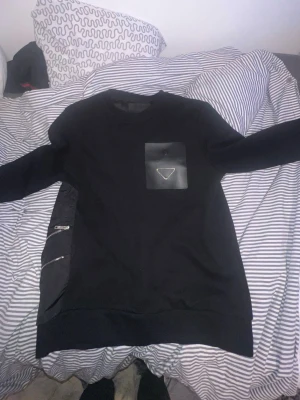 Svart sweatshirt från Prada - Säljer en svart sweatshirt från Prada med en unik bröstficka i nylon och den klassiska trekantsloggan. Tröjan har rund halsringning, långa ärmar och coola dragkedjedetaljer på sidan. Materialet är en mix av bomull och nylon för en lyxig känsla.