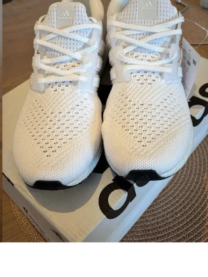 Vita Adidas Ultraboost sneakers - Snygga vita Adidas Ultraboost sneakers med stickad ovandel i mesh för bra andningsförmåga. Skorna har vita snören, klassiska tre ränder på sidan och en bekväm Boost-sula. Perfekta för dig som gillar stilrena och sportiga sneakers.