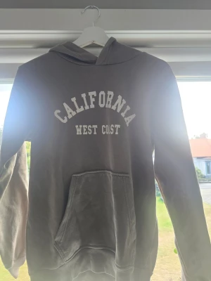 Brun hoodie California West Coast - Säljer en brun hoodie från Kappahl med texten 'CALIFORNIA WEST COAST' i vitt på bröstet. Hoodien har huva och en stor magficka framtill. Tillverkad i mjukt bomullsmaterial, perfekt för chill dagar och streetwear-vibe. Är I storleken 158/164 cm