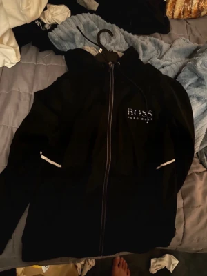 Svart hoodie från BOSS Hugo Boss - Säljer två svarta hoodies från BOSS Hugo Boss med en som har dragkedja framtill och huva med snören. Regular fit-modell med grå insida i halsen och vita detaljer på ärmarna och en hoodie som är också grå insidan.  Perfekt för en avslappnad och stilren look. Båda är storlek M!