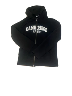 Sip-up hoodie från SOC - Svart hoodie med dragkedja och vit text 'Cambridge 1989' på bröstet. 
