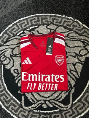 Arsenal matchtröja röd/vit Adidas - Snygg Arsenal matchtröja från Adidas i rött med vita ärmar och detaljer. Tröjan har klubbmärke och Adidas-logga på bröstet samt sponsortryck 'Emirates Fly Better' framtill. Tillverkad i lätt och ventilerande material, perfekt för fotboll eller träning.