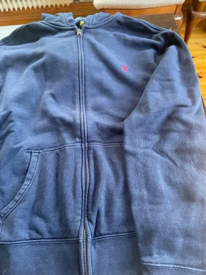 Mörkblå hoodie med dragkedja - Mörkblå hoodie med dragkedja framtill och fickor på sidorna. Klassisk röd broderad logga på bröstet. Tillverkad i mjukt bomullsmaterial och har en huva för extra komfort. Perfekt för en avslappnad och stilren look. Storlek XL i barn