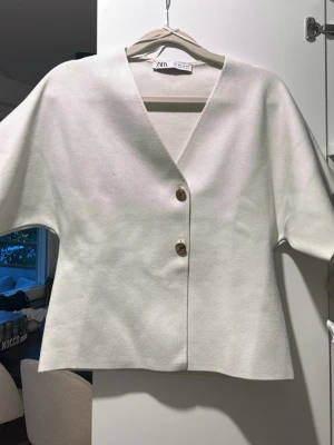 Vit kofta med knappar från Zara - Stilren vit kofta från Zara med v-ringning och två stora bruna knappar framtill. Koftan har vida, korta ärmar och rak passform. Perfekt att slänga över en topp för en clean look.