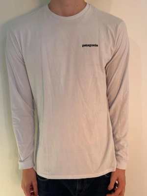 Vit långärmad Patagonia t-shirt - Vit långärmad t-shirt från Patagonia med liten logga på bröstet och stor färgglad logga med bergmotiv på ryggen. Tillverkad i mjuk bomull och har en klassisk passform med rund hals. Perfekt för en avslappnad och stilren look.