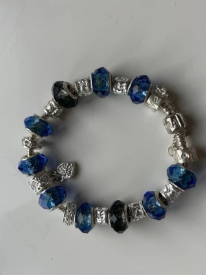 Pandora armband med blå pärlor - Säljer ett snyggt Pandora armband med silverfärgad kedja och flera blå  pärlor. Armbandet har detaljerade silverberlocker och är perfekt för dig som gillar att mixa och matcha smycken. 😊