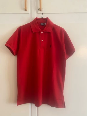 Polo Ralph Lauren röd pikétröja - Klassisk Polo Ralph Lauren röd pikétröja med korta ärmar och krage. Tröjan har två knappar framtill och en liten broderad logga på bröstet. Perfekt till jeans eller shorts för en enkel och snygg look.