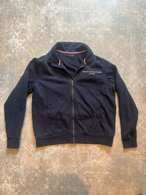 Marinblå ziptröja Tommy Hilfiger - Snygg marinblå ziptröja från Tommy Hilfiger med hög krage och dragkedja framtill. Tröjan har två fickor, broderad logga på bröstet och detaljer i rött och vitt på insidan av kragen. Perfekt för en avslappnad och stilren look.