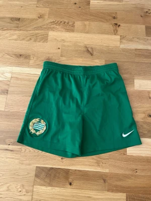 Gröna Hammarby-shorts från Nike - Gröna sportshorts från Nike med elastisk midja och broderad Nike-logga i vitt på ena benet. Shortsen har även en snygg broderad emblem-detalj i guld och grönt på andra benet. Perfekta för träning eller sportiga dagar.