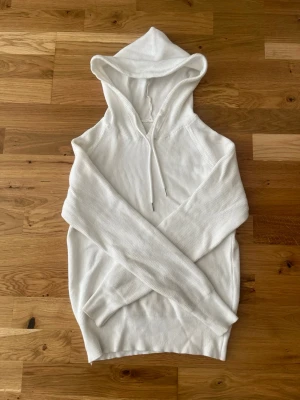 Vit stickad hoodie - Säljer en stilren vit hoodie i stickat material med huva och dragsko. Hoodien har långa ärmar och breda muddar vid ärmslut och nederkant. Perfekt för en avslappnad och trendig look.