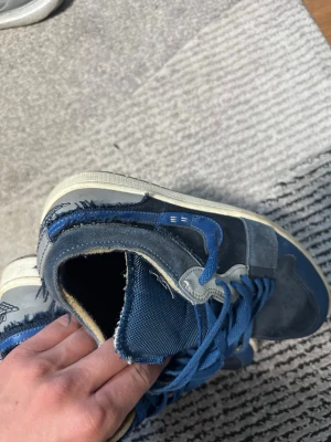 Blå höga sneakers med vita detaljer - Snygga höga sneakers i olika blå nyanser med vita detaljer och blå skosnören. Ovandelen är i en mix av mocka och textil, med perforerad tåbox och en ikonisk logga på plösen. Sulan är vit med blå inslag. Perfekta för dig som gillar streetwear.