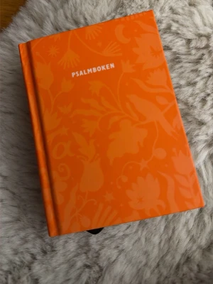 Psalmboken i orange hårdband - En svensk psalmbok med hårda pärmar i en stark orange färg och blommigt mönster. Boken innehåller både noter och text till psalmer. Perfekt för dig som gillar musik, sång och traditionella texter.