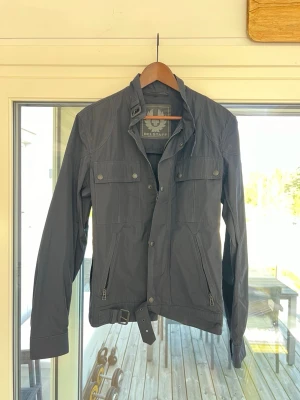 Svart vindjacka från Belstaff - Snygg svart vindjacka från Belstaff med ståkrage, bälte i midjan och flera fickor med knappar och dragkedjor. Jackan har långa ärmar och en klassisk, stilren look som passar perfekt till vardagsoutfiten.