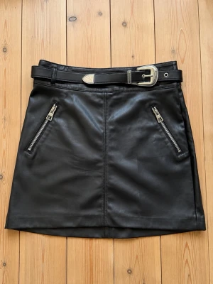 Svart kort kjol i skinnimitation - Snygg svart kort kjol i skinnimitation med två dragkedjefickor framtill och ett brett bälte med silverdetaljer. Kjolen har en rak passform och är perfekt för att skapa en edgy look.