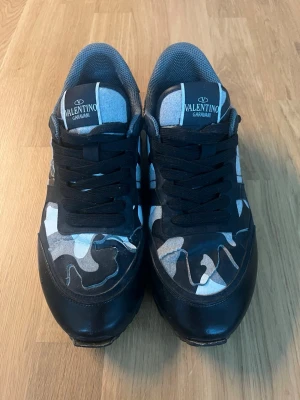 Valentino Garavani camo sneakers - Snygga sneakers från Valentino Garavani med svart, grå och vit camo-mönstrad ovandel. Skorna har svarta skosnören, meshdetaljer och robust gummisula. Perfekta för dig som vill sticka ut med en unik och trendig look.