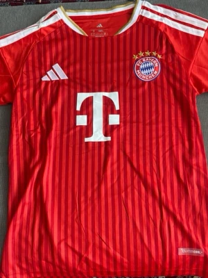 Röd FC Bayern München matchtröja - Säljer en röd FC Bayern München matchtröja från Adidas med vita detaljer och smala vertikala ränder. Tröjan har klubbmärke på bröstet, Adidas-logga och stor vit T-logga på framsidan. Tillverkad i lätt och ventilerande material med Climacool-teknologi.