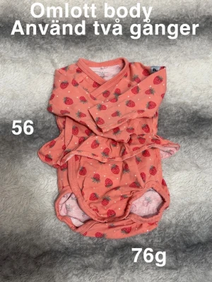 Rosa omlottbody med jordgubbar - Supersöt omlottbody i rosa med jordgubbsmönster och små vita prickar. Långa ärmar och praktisk omlottknäppning gör den enkel att ta på och av. Perfekt för dig som gillar gulliga och färgglada plagg till de minsta.🌸 Använd två gånger. Storlek 56 