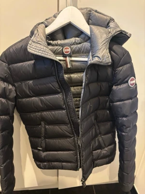Svart dunjacka från Colmar med huva - Snygg svart dunjacka från Colmar med quiltad design och fast huva. Jackan har dragkedja framtill, två sidofickor och Colmar-logga på bröstet och ärmen. Perfekt för kalla dagar och ger en sportig look.