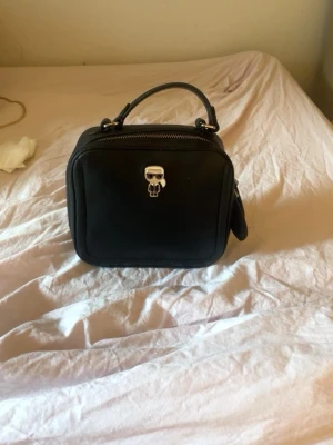 Svart handväska från Karl Lagerfeld - Snygg svart handväska i boxig form från Karl Lagerfeld-emblem på framsidan. Väskan har ett kort handtag upptill och dragkedja. Perfekt accessoar för dig som gillar stilrena detaljer och ikoniska designer.