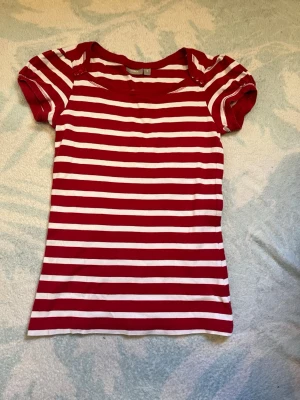 Rödvit randig t-shirt - Säljer en röd och vit randig t-shirt med korta ärmar. T-shirten har en klassisk rund halsringning och är gjord i mjuk bomull, perfekt för en avslappnad stil. Passformen är normal och tyget känns skönt mot huden.