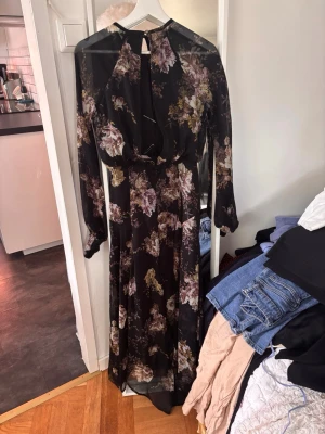 Blommig långklänning med nyckelhålsringning - Säljer en svart långklänning med blommigt mönster i lila, beige och gröna toner. Klänningen har lång ärm, markerad midja och en snygg nyckelhålsringning fram. Tunt, flytande material som ger en elegant look.
