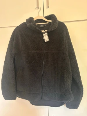 Svart teddyjacka med dragkedja - Mysig svart teddyjacka med hög krage och dragkedja framtill. Jackan har en fluffig, mjuk yta och två sidofickor. Perfekt för kyliga dagar och ger en avslappnad look. Materialet är typiskt teddy/fleece som håller värmen bra.