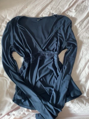 Mörkblå omlottopp från Gina Tricot - Snygg mörkblå omlottopp från Gina Tricot med spetsdetalj i midjan. Toppen har lång ärm och v-ringad halsringning, vilket ger en stilren och trendig look. Perfekt att matcha med jeans eller kjol. Jag har även samma tröja i vinröd!