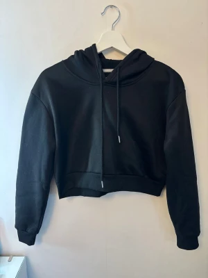Svart cropped hoodie med tryck - Svart cropped hoodie med huva och dragsnören. Tillverkad i mjukt sweatshirtmaterial, perfekt för en avslappnad streetwear-look.