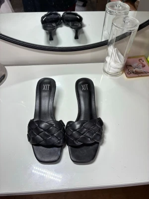 Svarta flätade pumps från XIT - Säljer ett par svarta pumps från XIT med trendig flätad rem över foten och öppen tå. Skorna har en låg klack och fyrkantig tå, vilket ger en modern look. Tillverkade i konstläder och perfekta för dig som gillar stilrena detaljer.