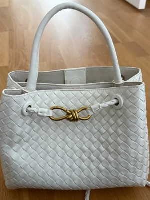 Vit flätad handväska med gulddetalj - Snygg vit handväska i flätat material med två handtag och dekorativ guldfärgad metallknut framtill. Väskan har en stilren och modern design med praktiska detaljer och rymligt fack. Perfekt accessoar för att lyfta din outfit.