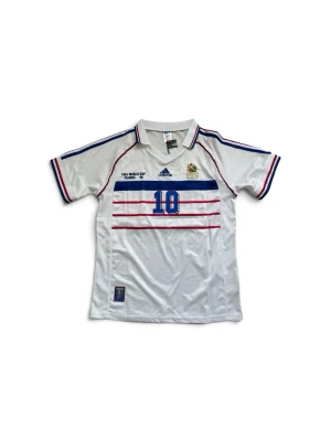 Frankrike fotbollströja Zidane #10 - Klassisk vit fotbollströja från Adidas med blå och röda detaljer, inspirerad av Frankrikes landslagströja från VM 1998. Nummer 10 och Zidane tryckt på ryggen, korta ärmar och krage. Perfekt för dig som älskar retro och fotboll. Kom gärna med bud!