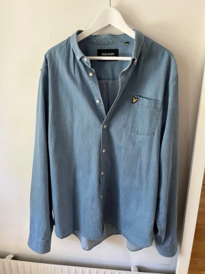 Ljusblå skjorta från Lyle & Scott - Säljer en stilren ljusblå skjorta från Lyle & Scott med klassisk krage och knappstängning. Skjortan har en bröstficka med den ikoniska gula loggan och är tillverkad i mjuk bomull. Perfekt till jeans eller chinos för en clean look.