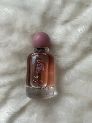Miss Laverni Bloom parfym - En elegant parfymflaska med rundad form och rosa tonad glas. Locket är matt rosa och flaskan har en stilren design med guldfärgad text och ett dekorativt mönster framtill. Perfekt för dig som gillar fräscha och blommiga dofter.