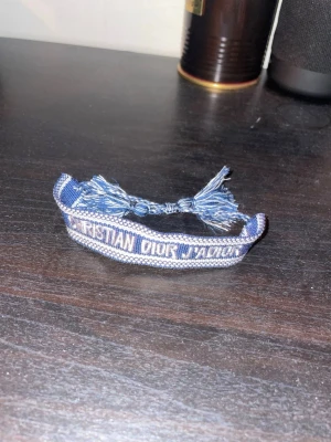 Blått Christian Dior J'Adior armband - Helt ny,Snyggt blått armband från Christian Dior med texten 'J'Adior' broderad i vitt. Armbandet är gjort i tyg och har justerbara knytband med fransar. Perfekt accessoar för att lyfta din outfit med en lyxig touch.