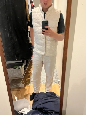 SAND VÄST NY - Stilren beige dunväst med raka paneler och knäppning framtill. Västen har en diskret bröstficka och står upp i kragen för en modern look. Perfekt att slänga över en hoodie eller tröja för extra värme. Passar till jeans och funkar året runt. Aldrig använd.