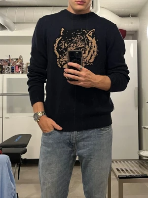Svart stickad tröja med tigertryck - Säljer en svart stickad tröja från Tiger of Sweden med ett stort beige tigerhuvud på bröstet. Tröjan har rund halsringning och ribbade muddar vid ärmslut och nederkant. Perfekt för dig som vill ha en cool och unik look.