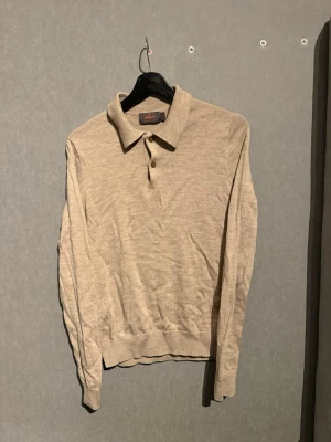 Morris pike långärmad - Stilren beige långärmad polotröja med klassisk krage och tre knappar framtill. Tröjan är tillverkad i ett tunt, mjukt material och har ribbade muddar vid ärmslut och nederkant. Perfekt för en clean och avslappnad look.