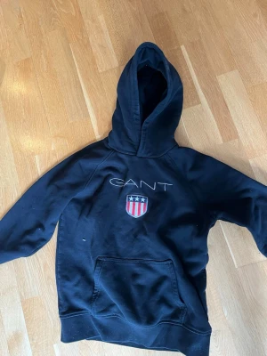 Svart hoodie från GANT med logga - Snygg svart hoodie från GANT med stor huva och klassisk logga broderad på bröstet tillsammans med ett sköldmärke i rött, vitt och blått. Hoodien har en stor magficka och ribbade muddar vid ärmar och nederkant. Perfekt för en avslappnad och cool stil. Strlk 146-152