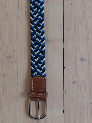 Flätat blått och brunt bälte - Snyggt flätat bälte i blå, vit och svart med bruna skinn-detaljer och silverfärgat spänne. 102 cm till spännet. 