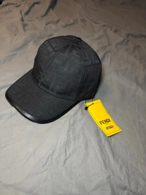 Svart keps från Fendi med logomönster - Snygg svart keps från Fendi med diskret ton-i-ton logomönster över hela kepsen. Klassisk böjd skärm och justerbar passform. Kepsen har en stilren look och är tillverkad i ett slitstarkt material. Perfekt accessoar för att lyfta din outfit.