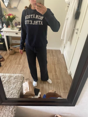 Mörkblå sweatshirt NONATION ATHLETIC - Mörkblå sweatshirt med trycket 'NONATION ATHLETIC' i vitt framtill. Tröjan har rund halsringning, ribbade muddar vid ärmslut och nederkant samt en avslappnad passform. Perfekt för en chill och sportig look.