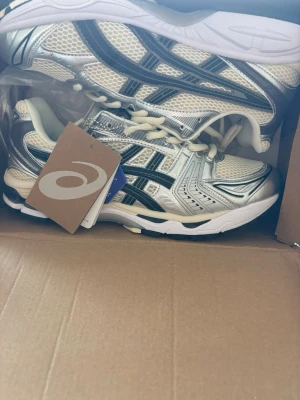 Vita och silverfärgade sneakers Asics - Snygga sneakers från Asics i vitt och silver med svarta detaljer. Ovandelen är i mesh och syntetmaterial för bra andningsförmåga och komfort. Klassisk låg modell med vita snören och robust sula. Perfekta för en sportig och trendig look.