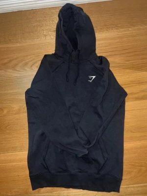 Svart hoodie från Gymshark - Säljer en svart hoodie från Gymshark med klassisk huva och dragsnören. Hoodien har en stor ficka framtill och Gymsharks logga i vitt på bröstet. Perfekt för chill dagar eller till gymmet. Materialet känns mjukt och bekvämt.