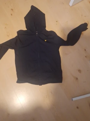Svart hoodie med dragkedja och logga - Säljer en svart hoodie med dragkedja och huva. På bröstet finns en liten gul broderad logga i form av en fågel. Hoodien har långa ärmar och fickor framtill. Perfekt för en chill och avslappnad stil.