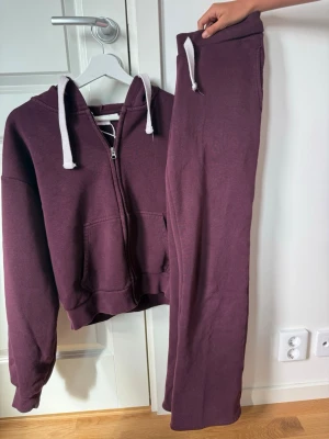 Vinröda mjukisbyxor med dragkedja  - Säljer ett vinrött mjukisset med zip-hoodie och matchande byxor. Hoodien har vita snören, dragkedja och fickor framtill. Byxorna har resår i midjan och vita snören. Båda delarna är i mjukt sweatshirtmaterial, perfekta för chill dagar.💕