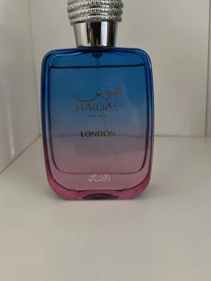 Hawas For Him London parfym - Snygg parfymflaska med blå till rosa toning och silverfärgad kork. Flaskan har en modern, fyrkantig form och text i guld på framsidan. Perfekt för dig som vill ha en unik och stilren doftaccessoar i badrumshyllan.