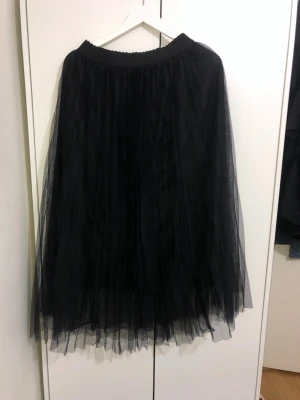 Svart långkjol i tyll - Säljer en svart långkjol i tyll med flera lager som ger ett fluffigt och voluminöst intryck. Kjolen har resår i midjan och ett elegant fall. Perfekt för dig som vill ha en statement piece i garderoben.