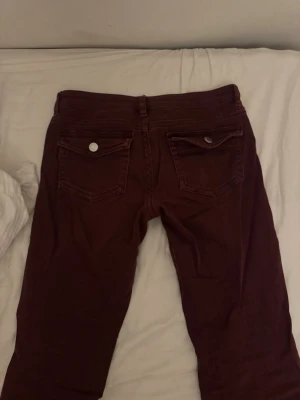 Vinröda jeans med bakfickor - Säljer ett par vinröda jeans med två bakfickor och silverfärgade knappar. Jeansen har normal midja och bälteshällor. Materialet är mjukt och stretchigt, vilket gör dem bekväma att bära. Perfekta för dig som vill ha ett par färgglada jeans i garderoben.