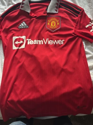 Manchester United matchtröja Sancho - Röd Manchester United matchtröja från Adidas med TeamViewer-tryck på bröstet och klubbmärke. Svarta och vita detaljer på axlar och krage. På ryggen står det 'Sancho' och nummer 25 i vitt. Tillverkad i lätt och ventilerande material, perfekt för fotboll.